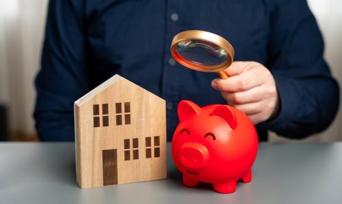 Person reviewing household finances with a piggy bank and model home while planning savings for unexpected costs.